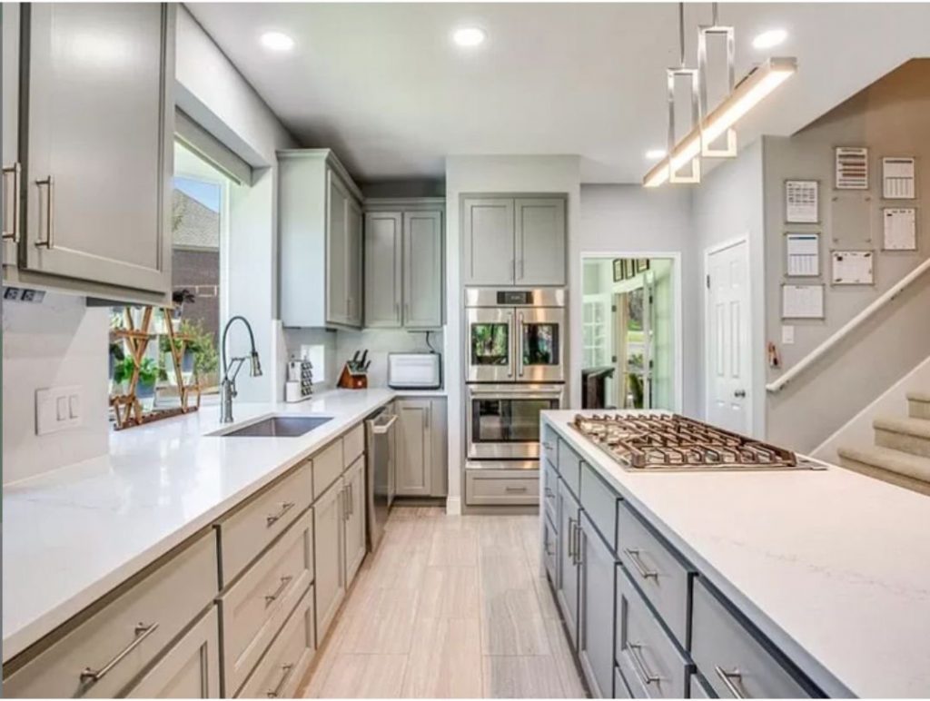 A modern kitchen featuring sleek gray cabinetry and pristine white quartz countertops. The kitchen includes a large central island with a built-in gas cooktop and contemporary pendant lighting overhead. The countertops offer ample workspace and a clean, elegant look. Stainless steel appliances, including a double oven and a built-in microwave, enhance the functionality and modern appeal of the space. A large window over the sink provides natural light, adding to the bright and airy atmosphere. The overall design showcases the beauty and practicality of high-quality kitchen countertops.