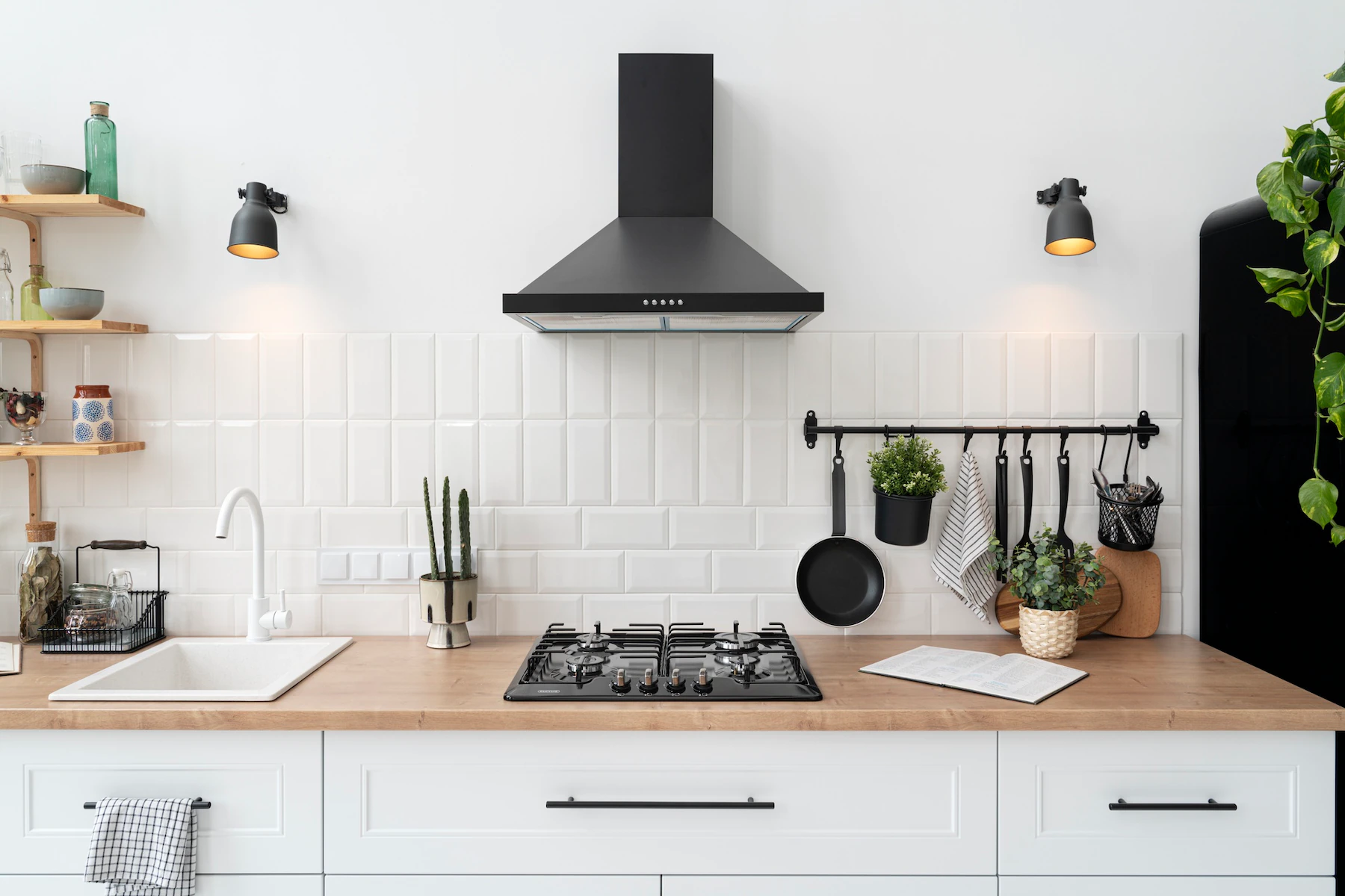 A stylish kitchen featuring a white subway tile backsplash with a slightly staggered pattern, providing a clean and modern look. The kitchen includes a black range hood above a gas stove, with a wooden countertop that adds warmth to the space. A white farmhouse sink is positioned to the left, and open wooden shelves hold various kitchen items. Black wall-mounted sconces provide additional lighting, and a black rail with hooks for hanging utensils and plants adds a practical yet decorative touch. This kitchen showcases a beautifully designed backsplash that complements the overall modern aesthetic.