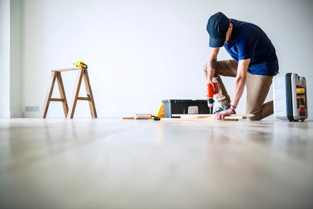 A professional installer working on a floor, using a power drill to secure wooden planks. The installer is dressed in a blue shirt and cap, and is kneeling on a light-colored hardwood floor. Nearby, a toolbox and various tools are visible, indicating an active flooring installation project. A wooden step ladder stands in the background, adding to the scene of a well-organized and precise flooring job. This image captures the expertise and attention to detail in floor installation.