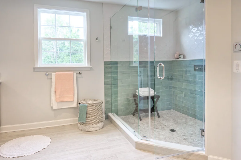 A beautifully remodeled guest bathroom featuring a spacious walk-in shower with clear glass doors and light blue subway tile walls. The bathroom includes a large window for natural light, a towel rack with a neatly folded peach towel, and a wicker basket for storage. The light wood flooring complements the serene and airy atmosphere, showcasing expert Kitchen & Bathroom Remodeling in Austin, TX.