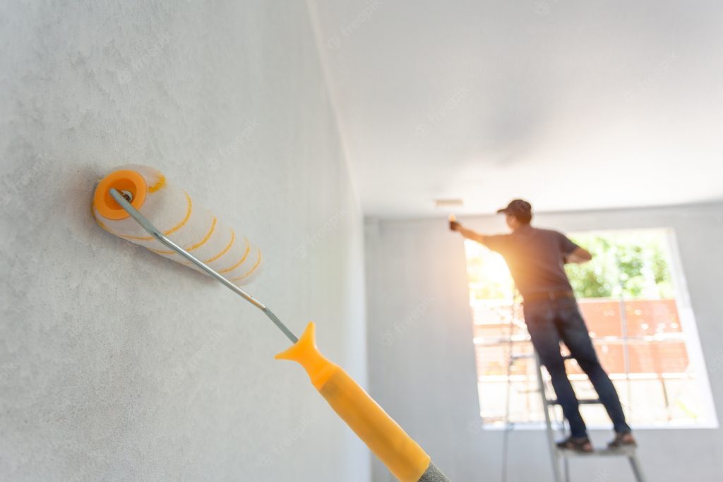 A close-up of a paint roller being used to apply a fresh coat of paint on a wall, highlighting the smooth and even coverage. In the background, a professional painter stands on a ladder near a window, working on additional painting tasks in a well-lit room. The sunlight streaming through the window creates a warm and inviting atmosphere, showcasing the attention to detail and quality of the painting services provided. This image captures the expertise and precision involved in professional painting services.