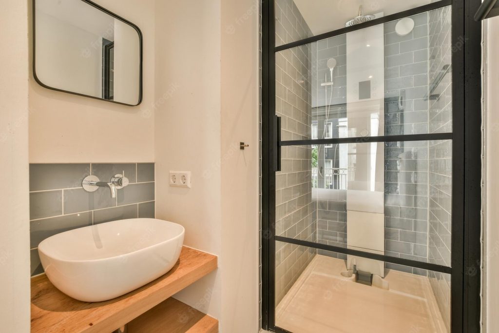 A modern shower remodel featuring a sleek, walk-in shower with gray subway tile walls and a black-framed glass door. Adjacent to the shower is a stylish wooden vanity with an integrated white vessel sink and a wall-mounted faucet, complemented by a minimalist mirror. The design emphasizes clean lines and a contemporary aesthetic, making the space both functional and visually appealing. This shower remodeling exemplifies high-quality craftsmanship and modern design.