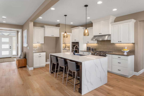 A beautifully remodeled kitchen featuring white cabinetry and a large island with a stunning waterfall marble countertop. The kitchen is equipped with stainless steel appliances, including a gas stove and range hood. Pendant lights hang above the island, which has seating for three. The backsplash is a sleek, light-colored tile that complements the neutral tones of the walls and cabinetry. Light wood flooring runs throughout the space, creating a warm and inviting atmosphere. This kitchen remodeling showcases a perfect blend of modern design and functionality.